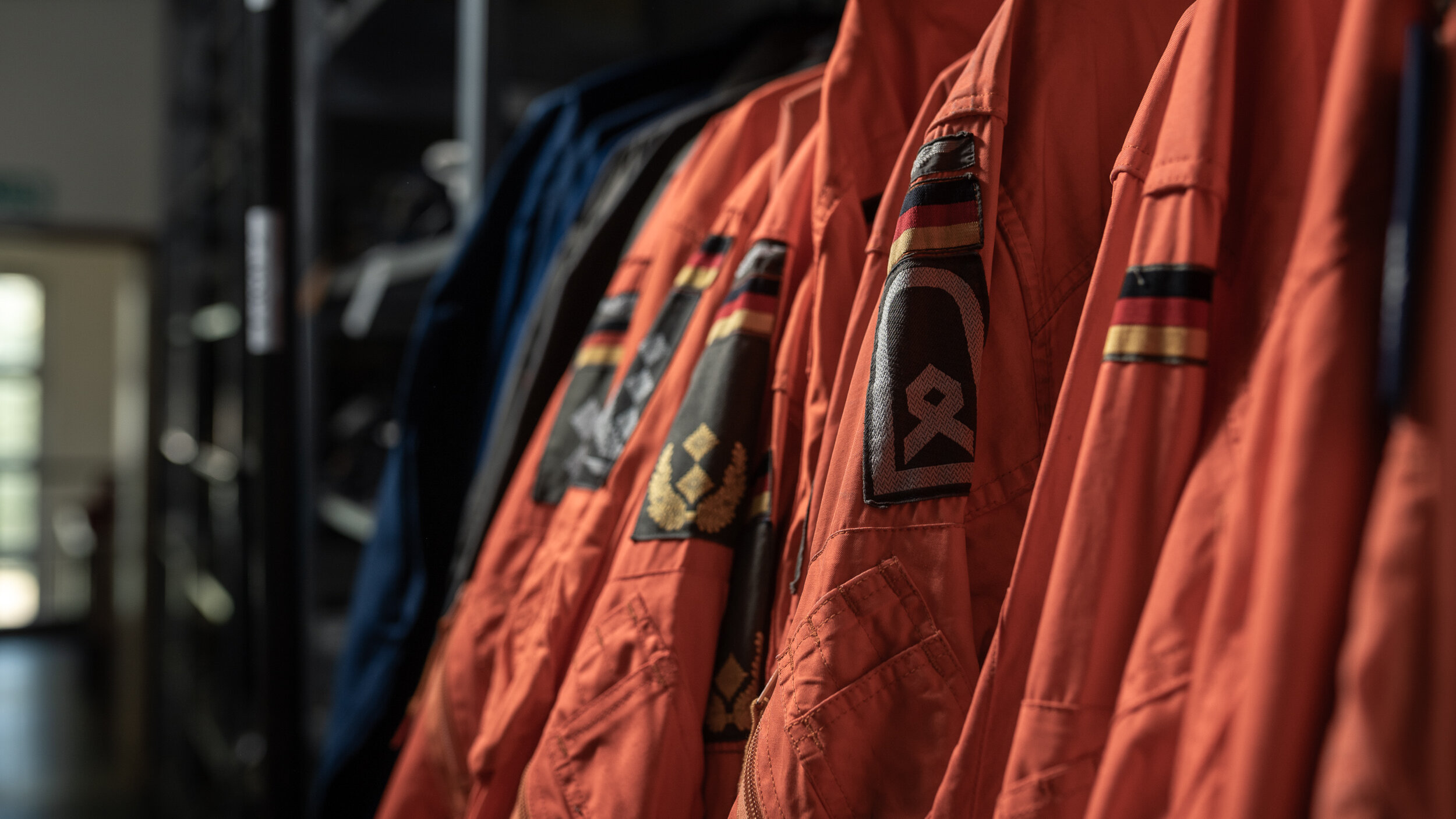 Orangerote Fliegerkombinationen mit Bundeswehr-Rangabzeichen hängen an einer Kleiderstange im Depot. 