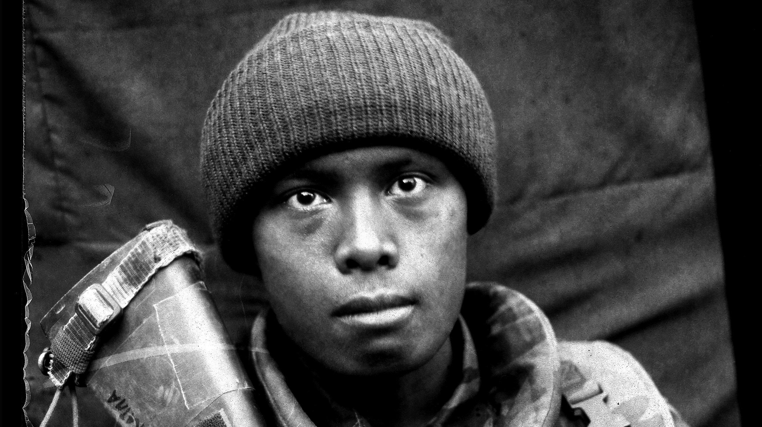 Schwarz-Weiß-Porträt eines jungen Soldaten, People of Color, in Uniform und mit Gewehr vor dunklem Hintergrund. 