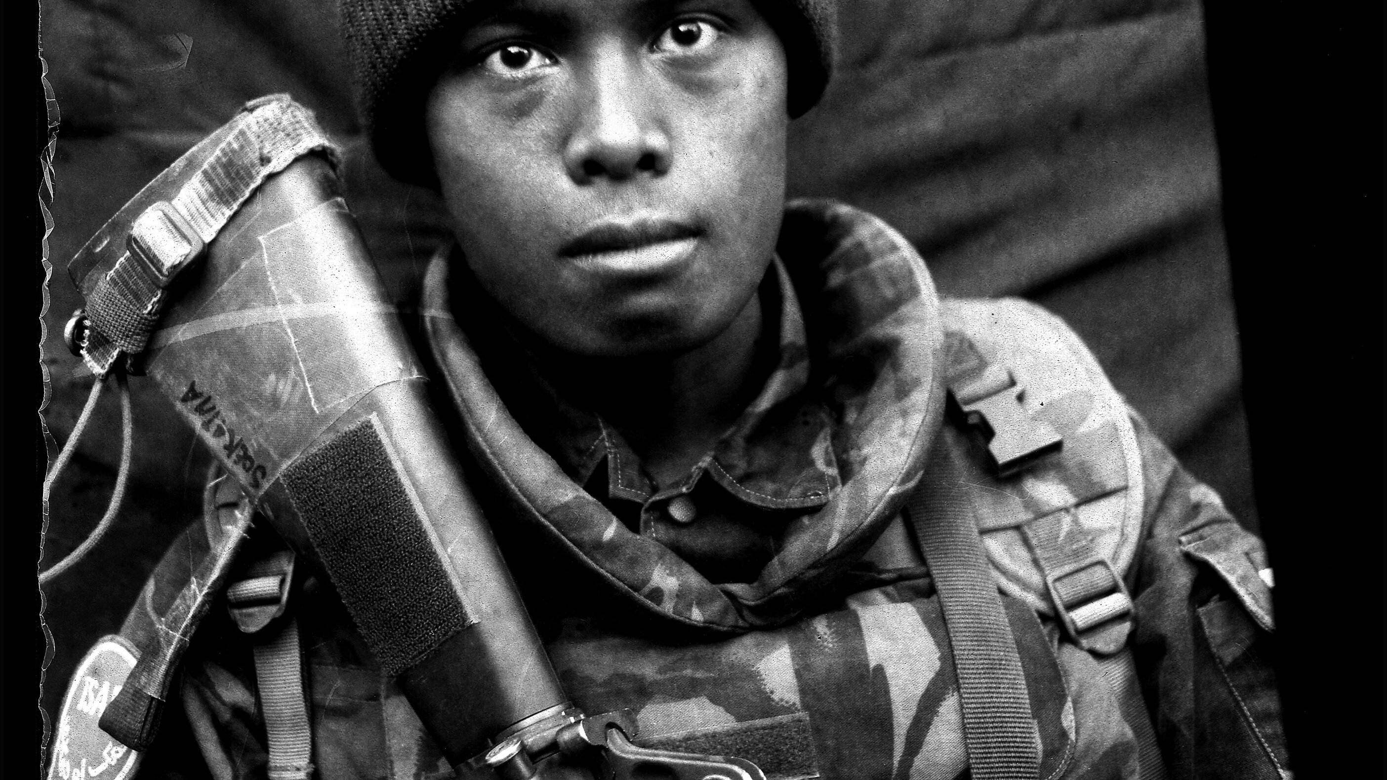 Schwarz-Weiß-Porträt eines jungen Soldaten, People of Color, in Uniform und mit Gewehr vor dunklem Hintergrund. 