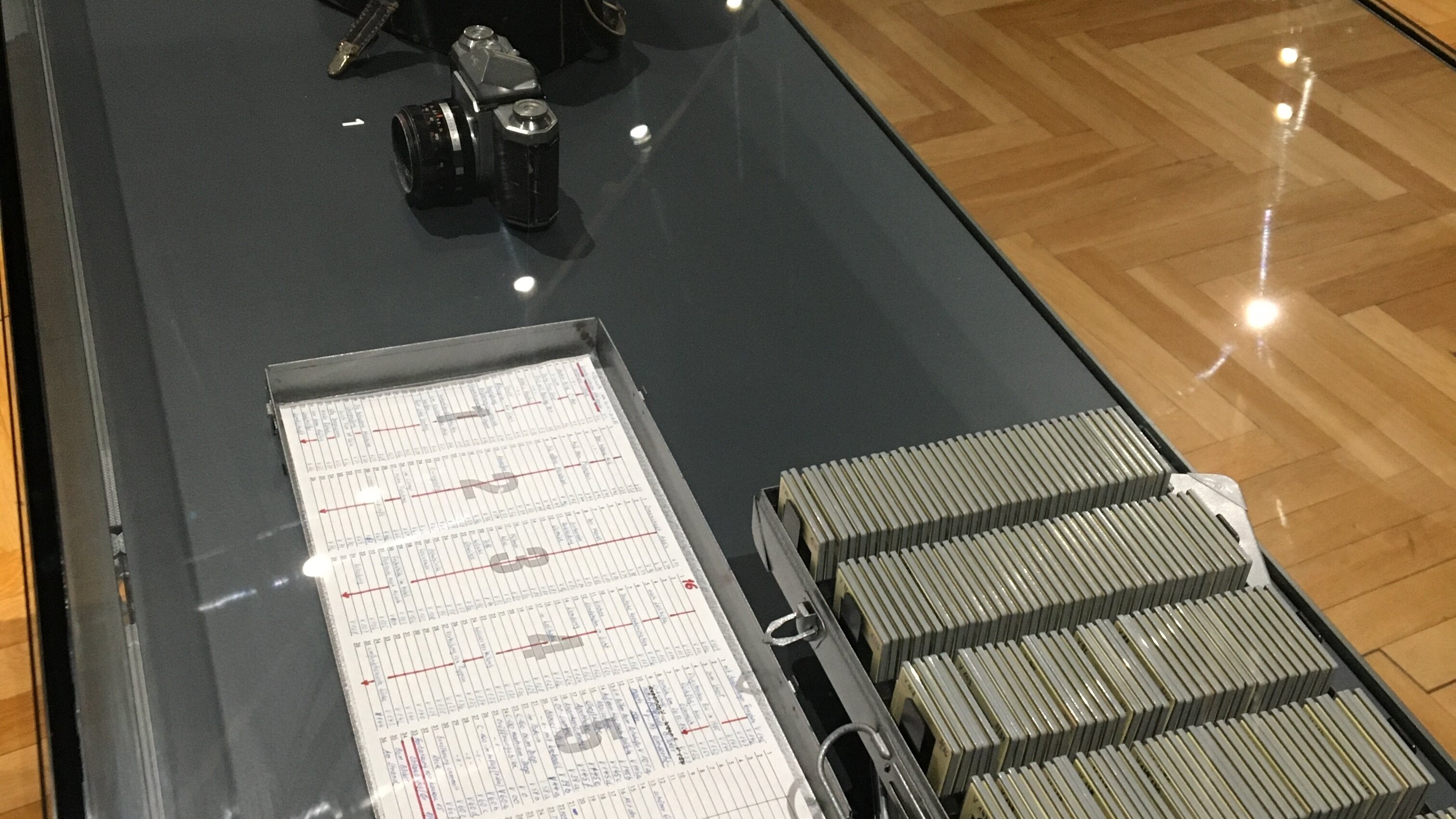 Blick in eine Vitrine mit gefülltem Diakasten, Kalender, Fotoapparat und Fototasche. 