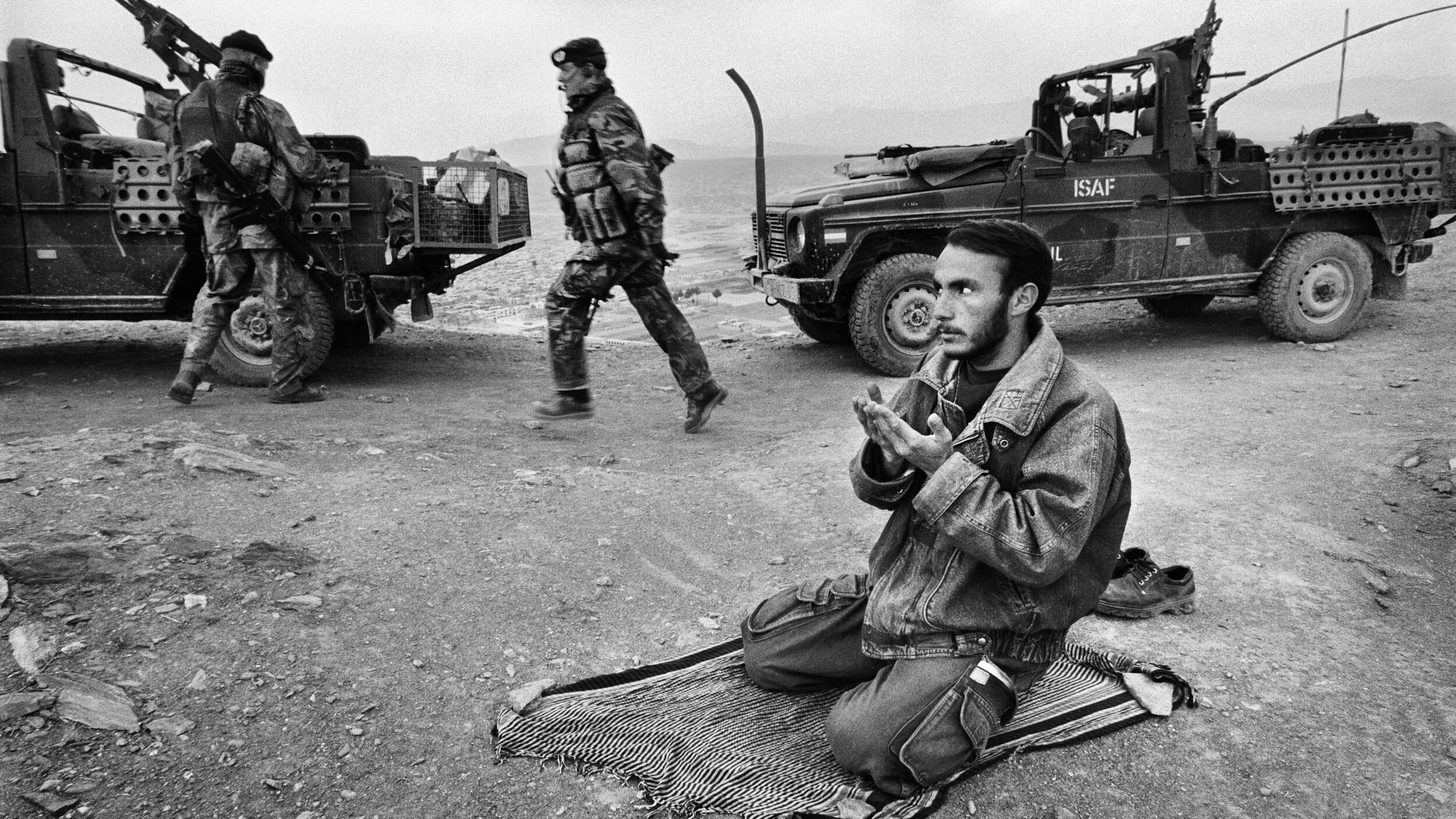 Ein junger afghanischer Mann kniet betend auf einer unbefestigten Straße, dahinter Militärfahrzeuge und Soldaten. 