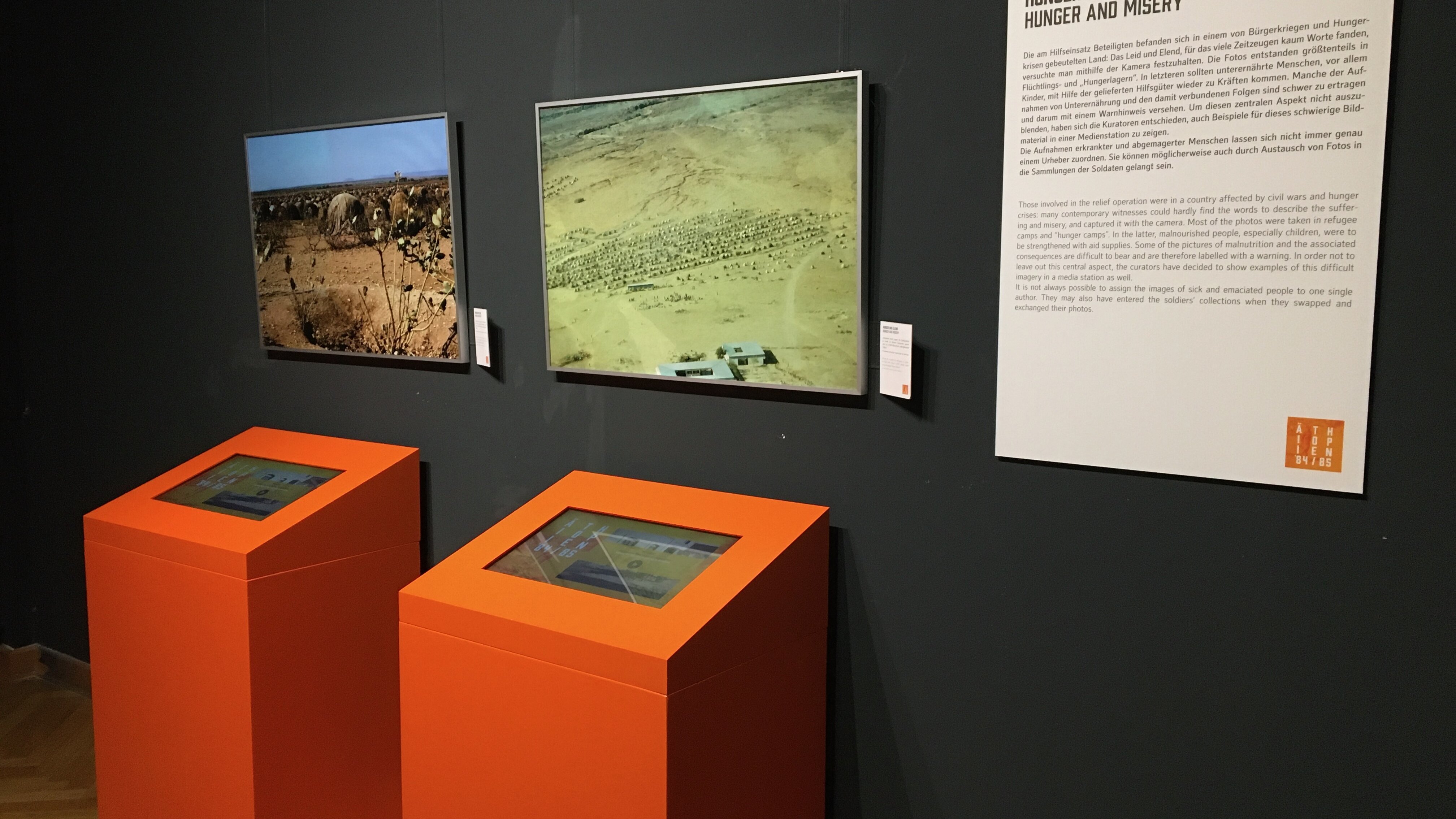 Schwarze Wand mit zwei Fotos, Texttafel und davor zwei orangefarbenen Medienstationen. 