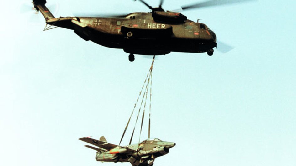 Ein kleines Jagdflugzeug hängt an einem großen Hubschrauber. 