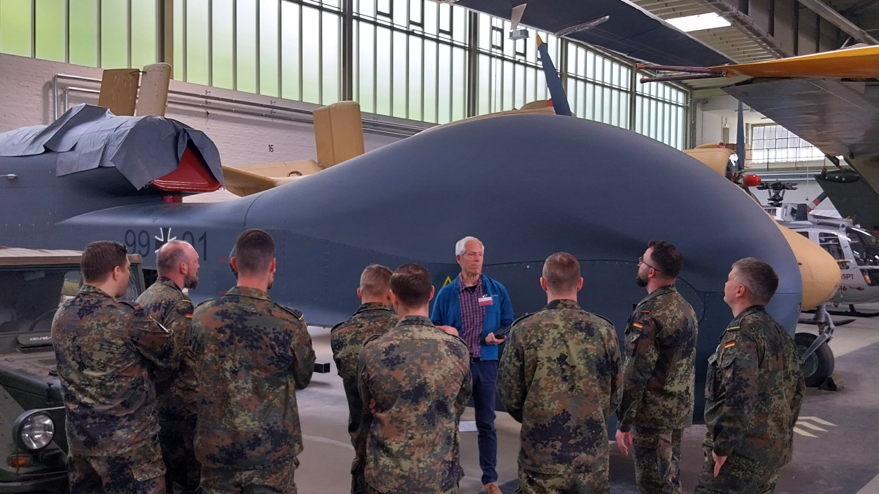 Eine Gruppe Soldaten im Feldanzug steht mit einem Guide vor der riesigen Drohne Euro Hawk im Depot. 