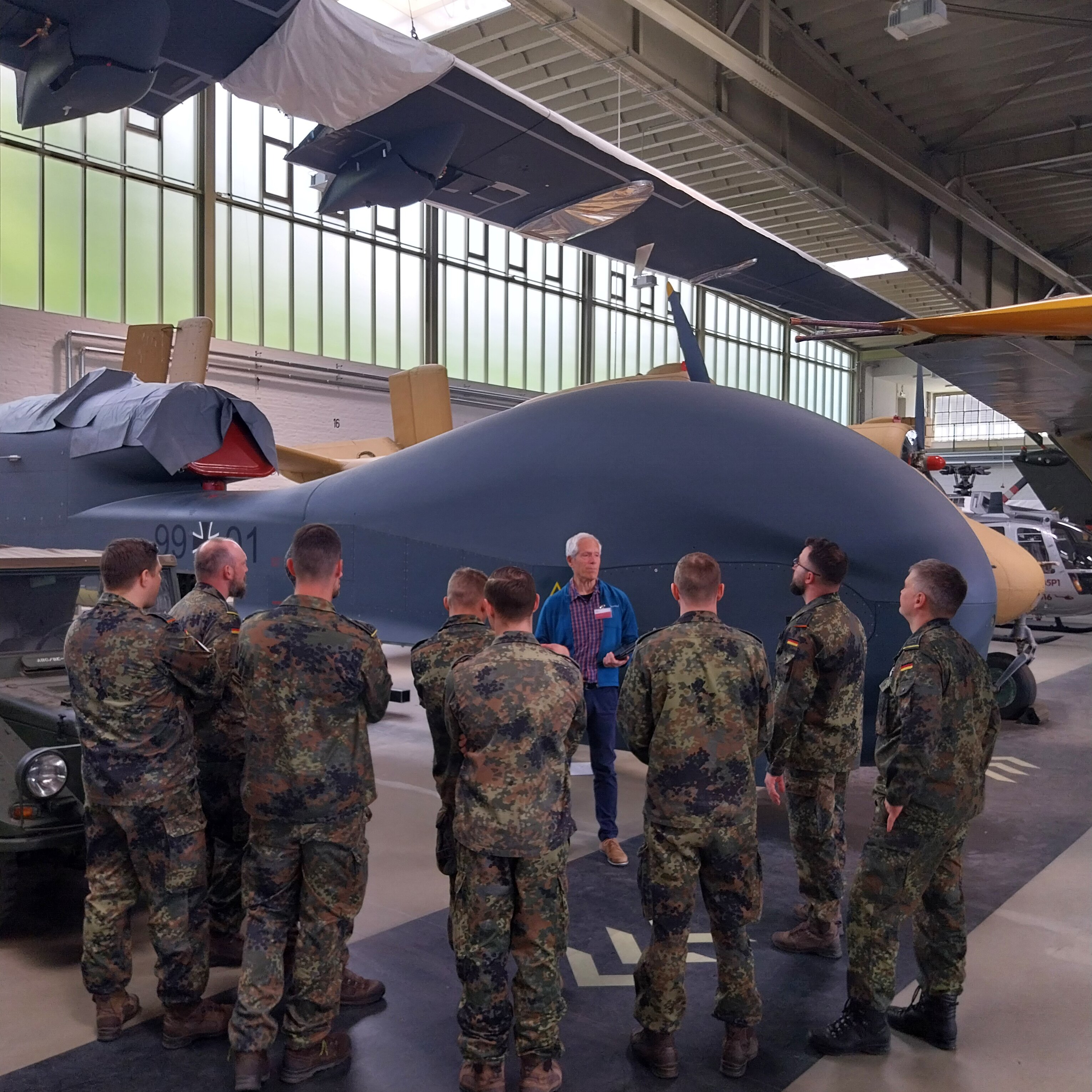 Eine Gruppe Soldaten im Feldanzug steht mit einem Guide vor der riesigen Drohne Euro Hawk im Depot. 