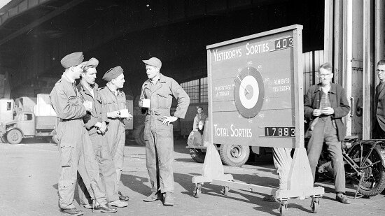 Vier britische Soldaten stehen neben einer Anzeigentafel vor einem offenen Hangar. 