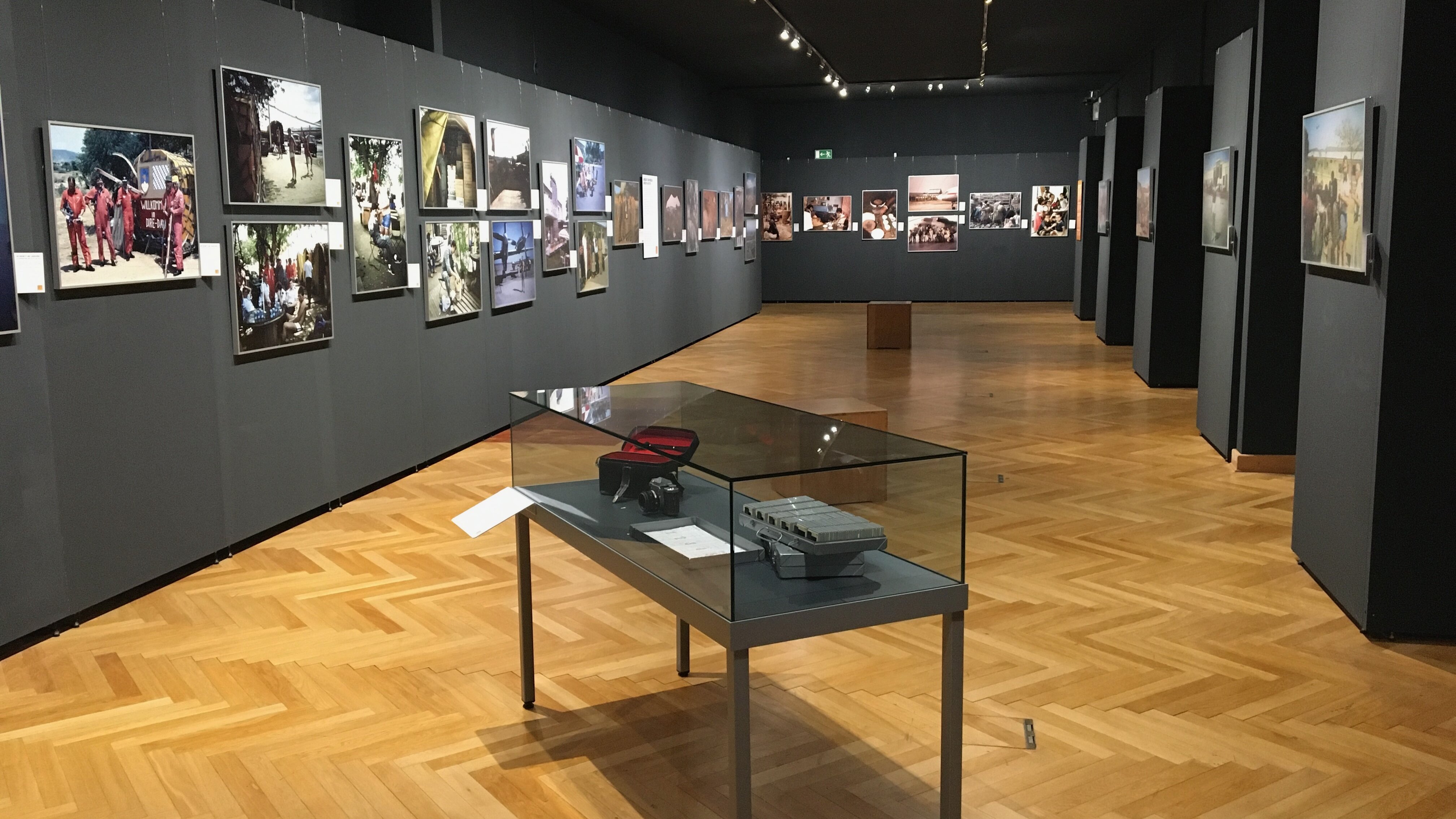Blick in den Ausstellungsraum mit Fotos an den dunklen Wänden und einer Vitrine im Vordergrund. 